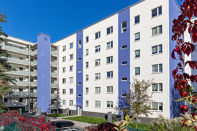 Modernisiertes Wohngebäude in Jena-Winzerla am Wacholderweg mit weiß-blauer Fassade, Balkonen und begrüntem Wohnumfeld.
